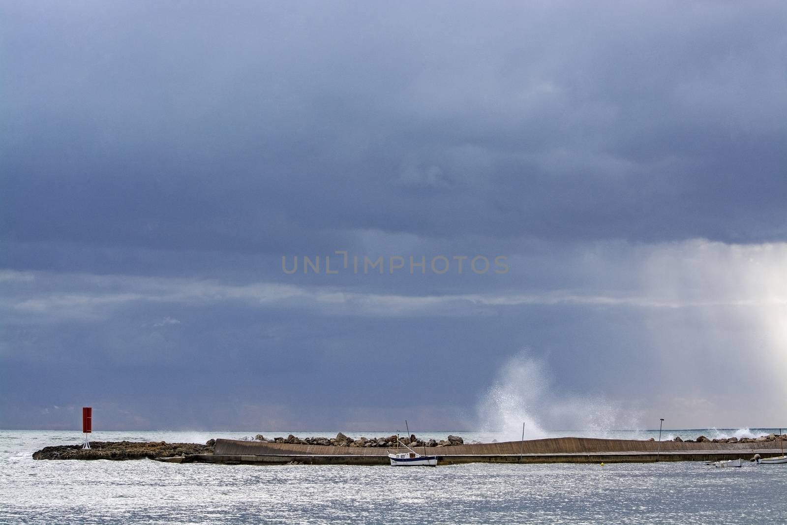 Mediterranean sea view with pier against dark skies with dramatic light on splashing wave on a winter day in Mallorca, Spain.