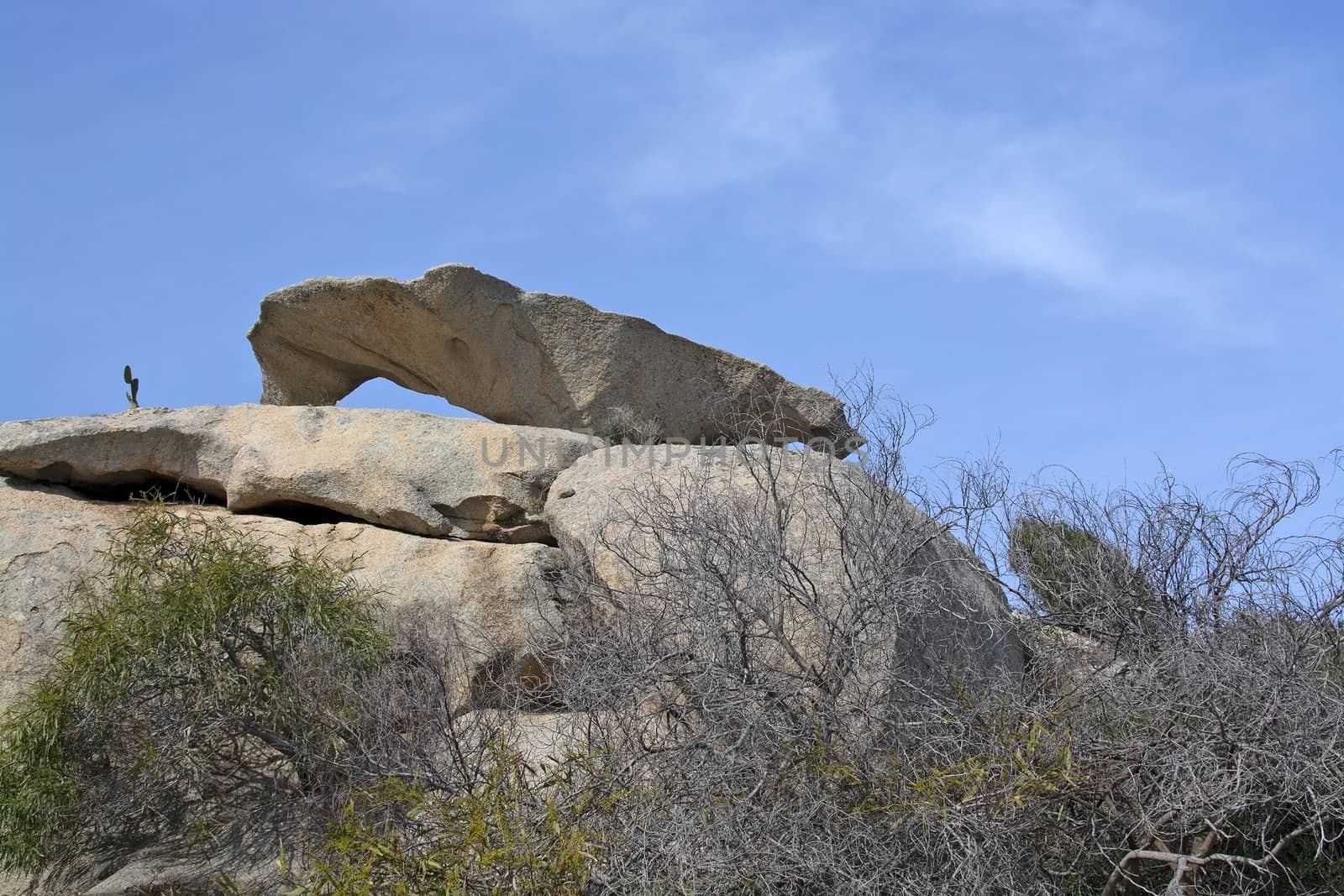 Slab of granite rock lying on top of eroded cliff with blue sky wispy cloud in Costa Smeralda, Sardinia, Italy.