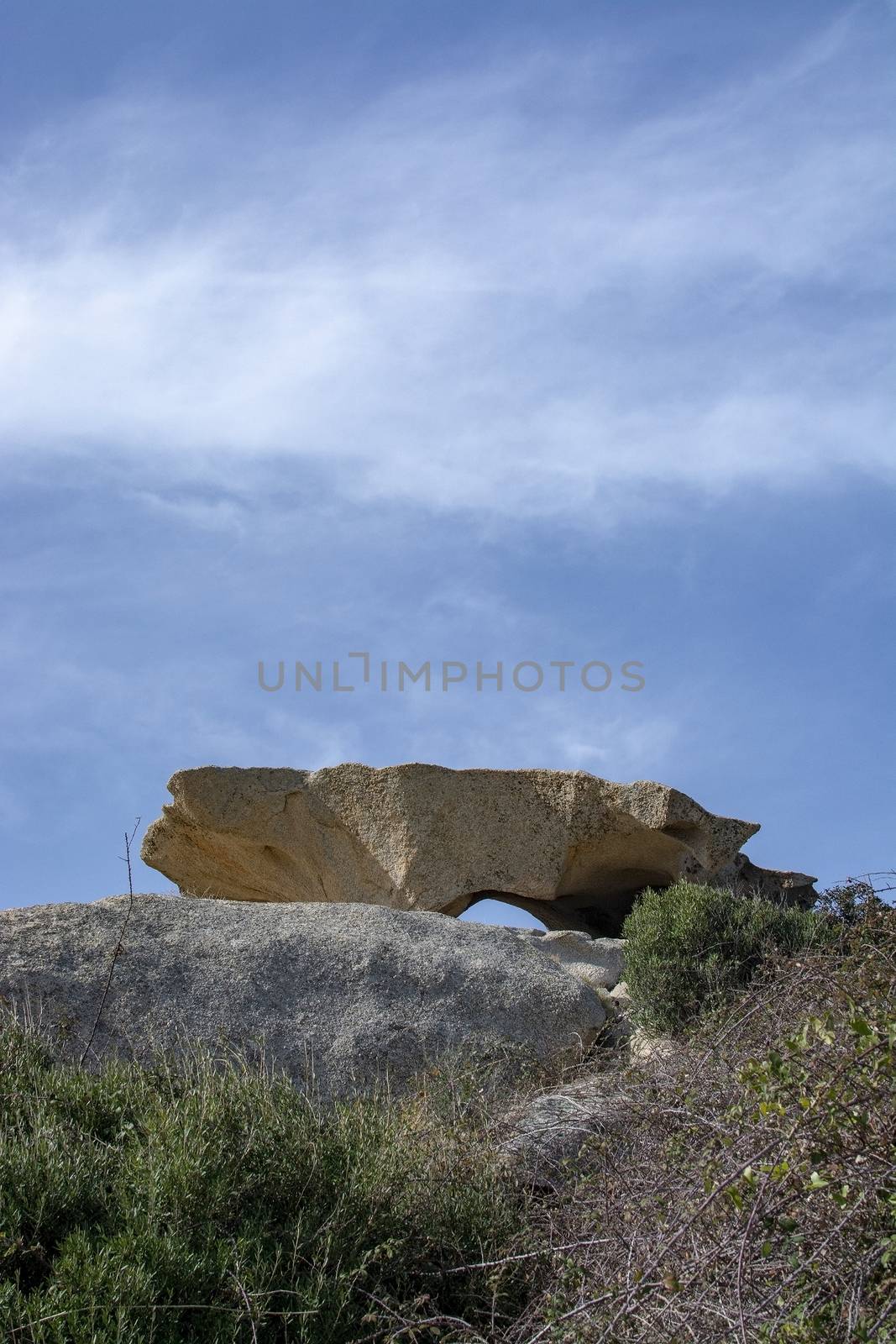 Slab of granite rock lying on top of eroded cliff with blue sky wispy cloud in Costa Smeralda, Sardinia, Italy.