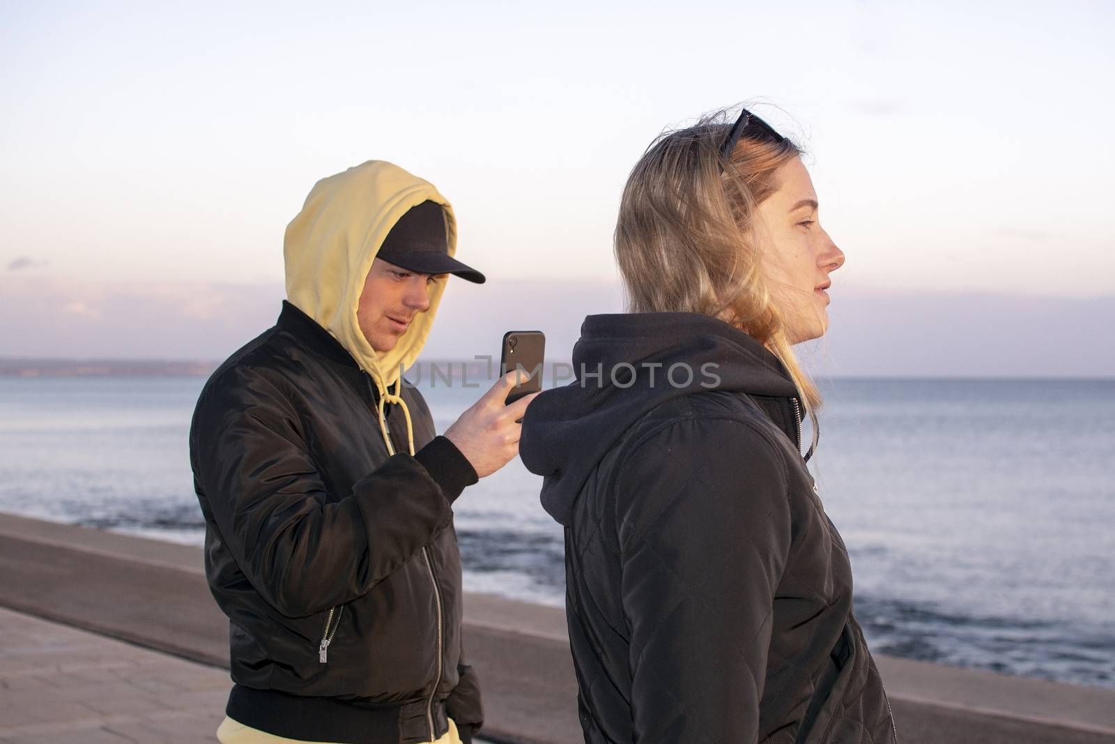 Handsome young natural and casual looking couple with hood jackets black and yellow watch sunset and photographing with smartphone on a beach.