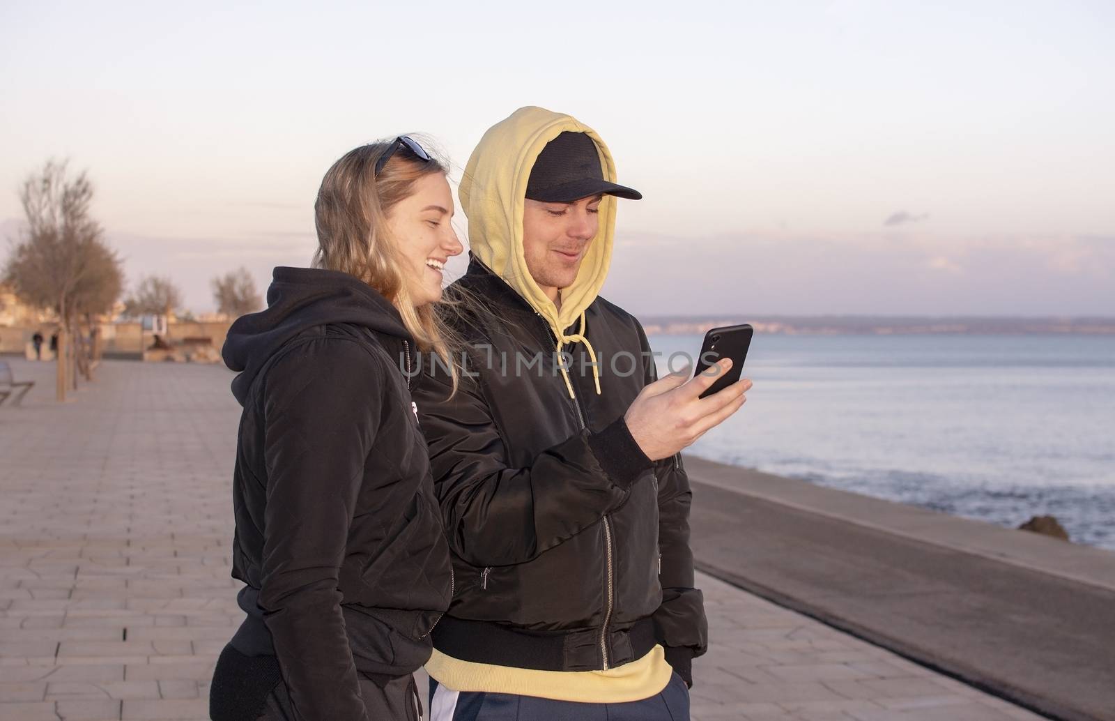 Handsome young natural and casual looking couple with hood jackets black and yellow watch sunset and photographing with smartphone on a beach.