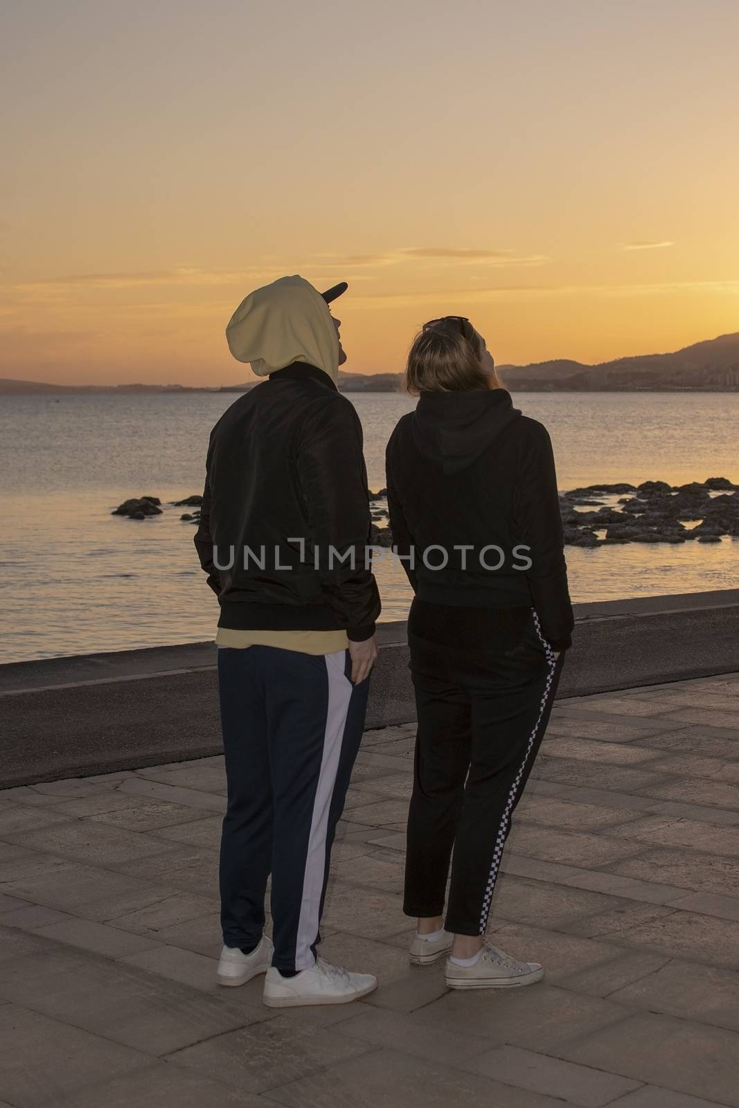 Vertical full body shot of handsome young natural and casual looking couple with hood jackets watch up towards sunset sky on a beach.