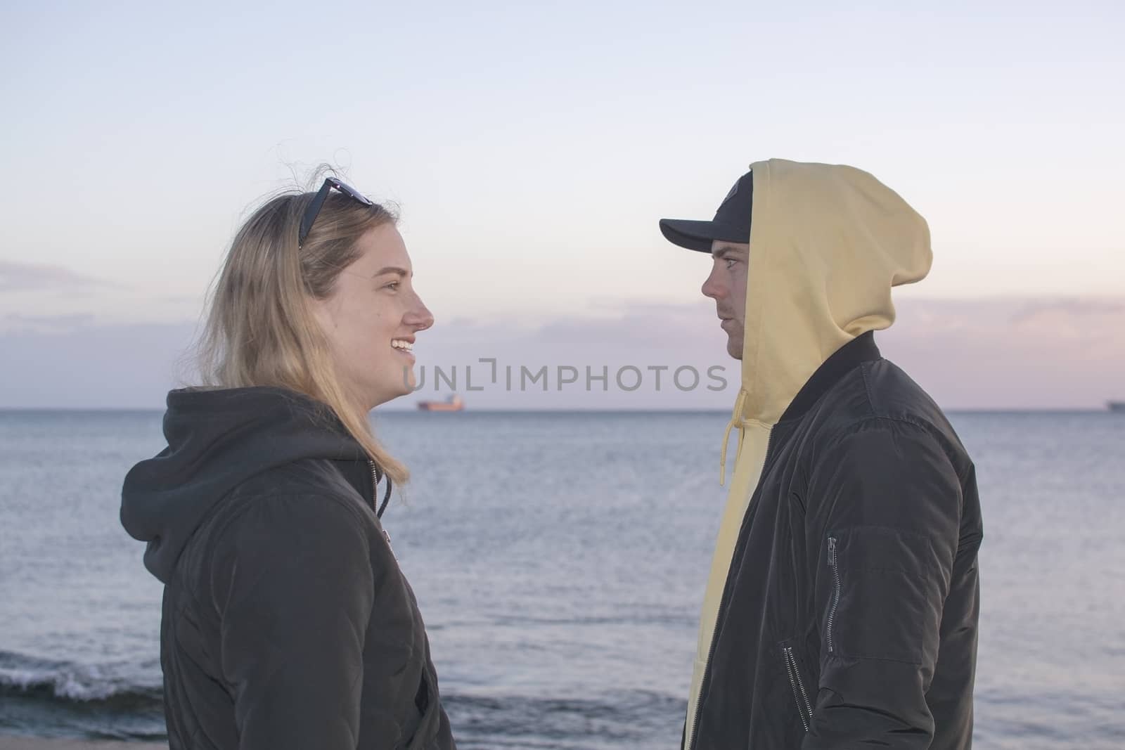 Handsome young natural and casual looking couple with hood jackets watch each other in profile at sunset on a beach.