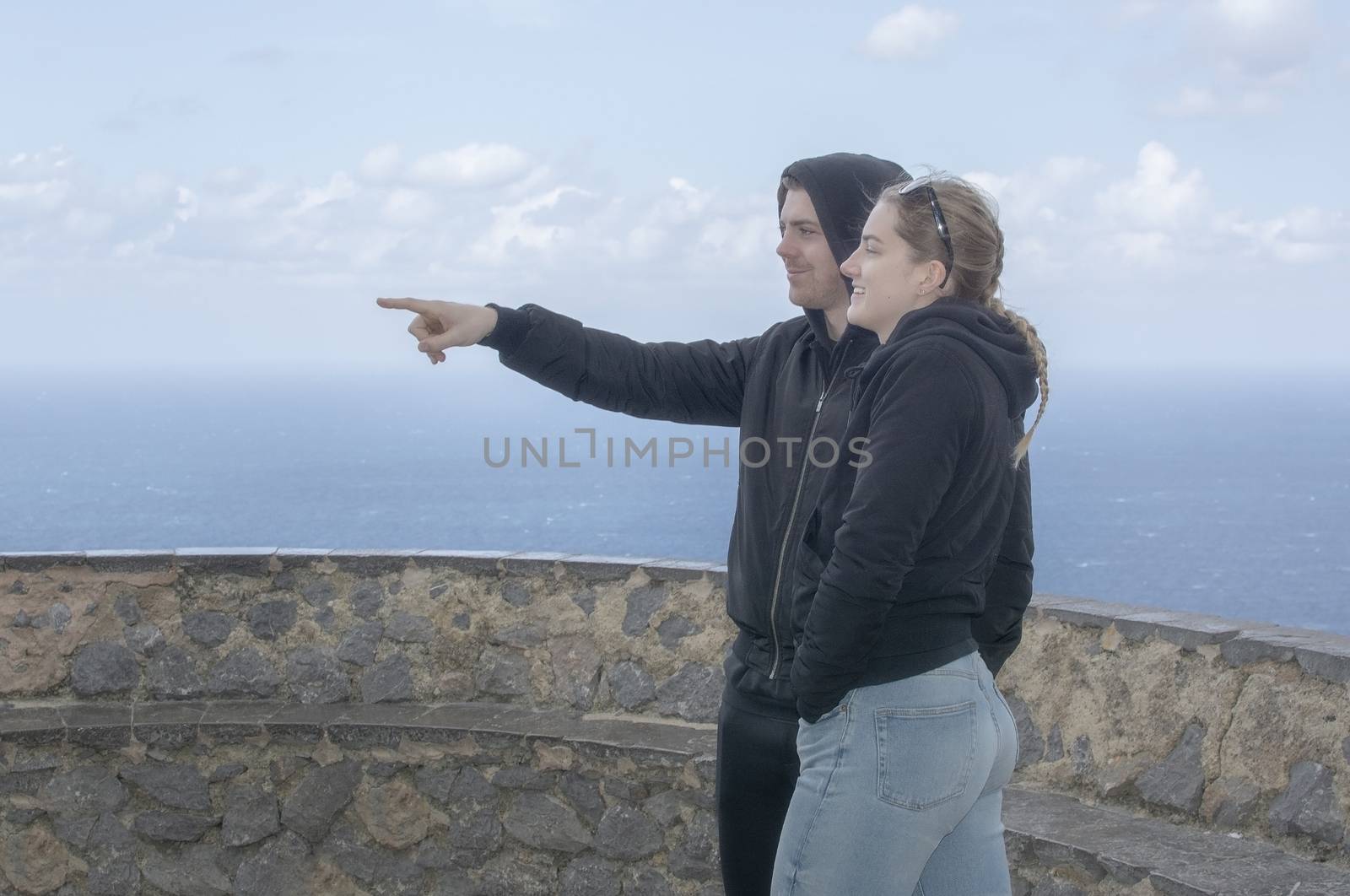 Young beautiful casually dressed couple stand high up pointing and watching out to sea on a spring day in Mallorca, Spain.