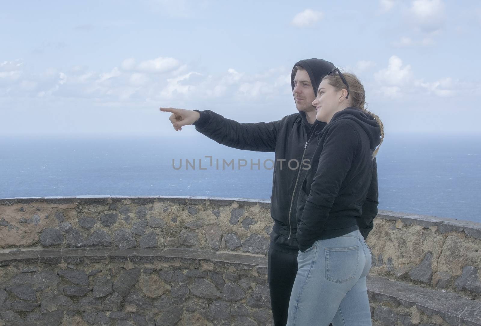 Young beautiful casually dressed couple stand high up pointing and watching out to sea on a spring day in Mallorca, Spain.