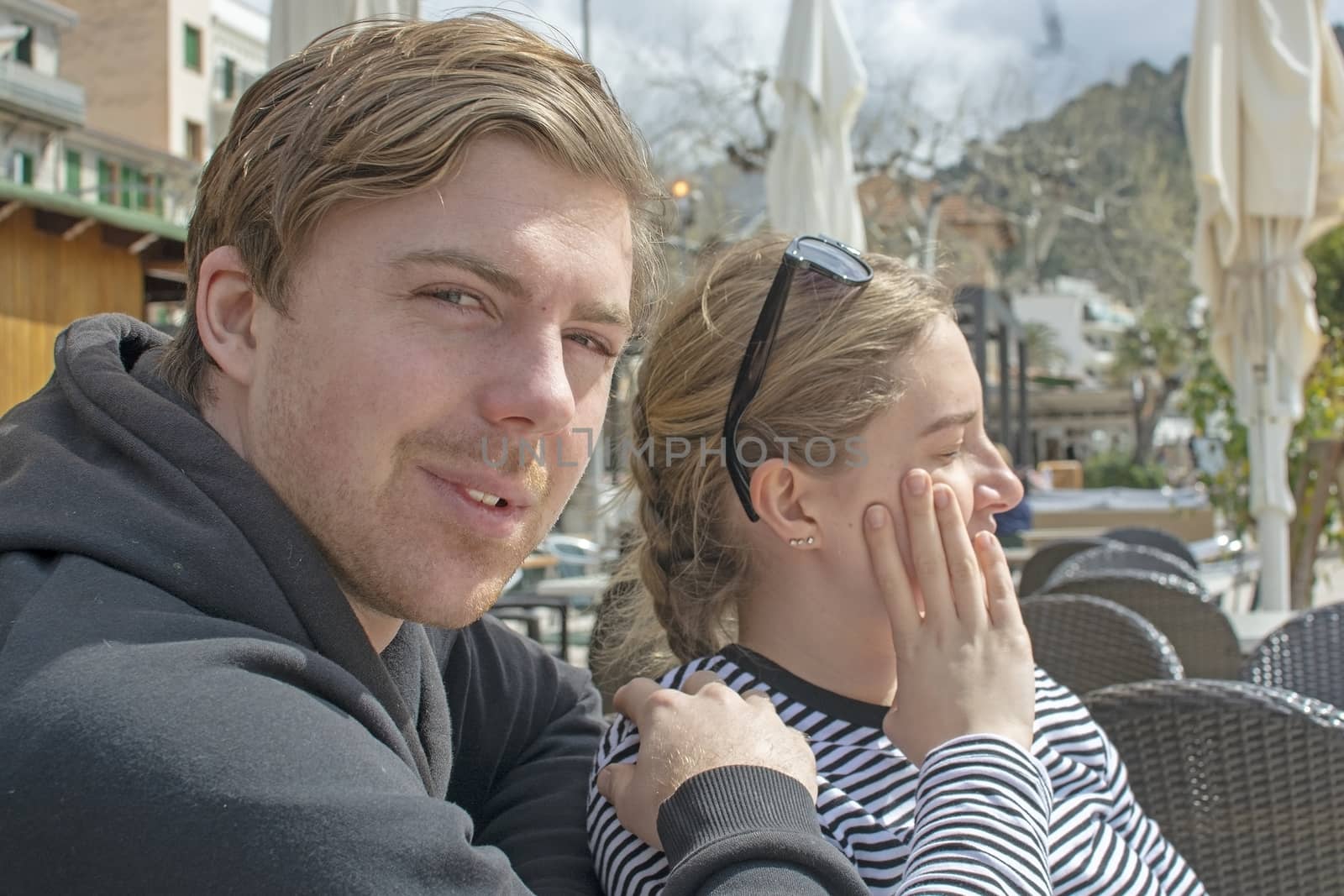 Young handsome happy couple sit close in cafe and squint in stark sunshine on a sunny spring day in Mallorca, Spain.