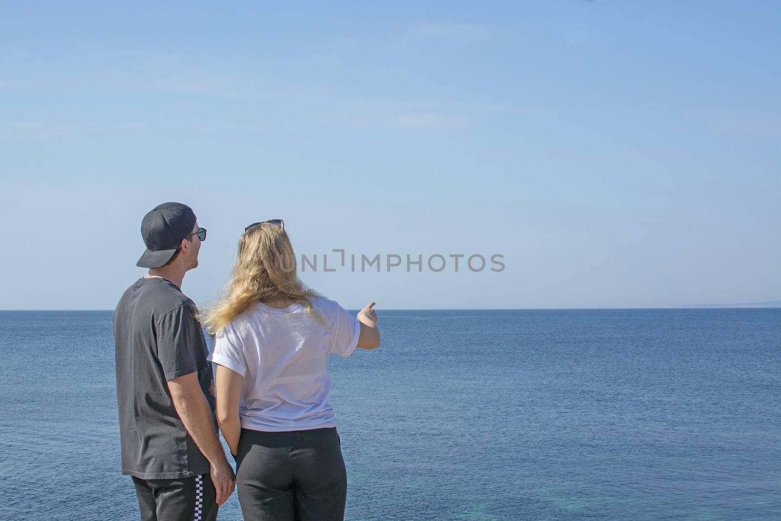 Young beautiful casually dressed couple stand high up pointing and watching out to sea on a spring day in Mallorca, Spain.