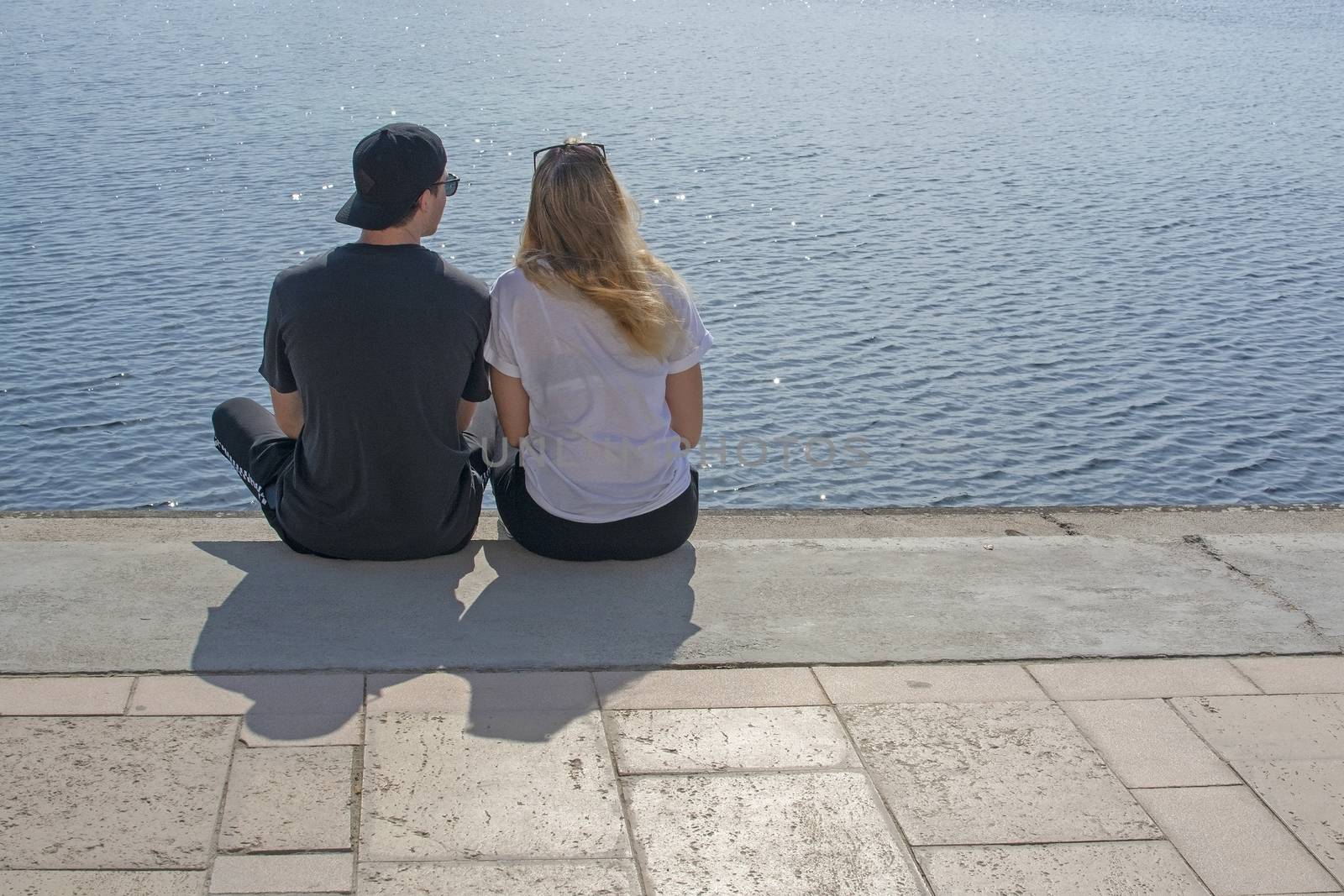 Young couple sit in sunshine watch water glitter on stone pier  by ArtesiaWells