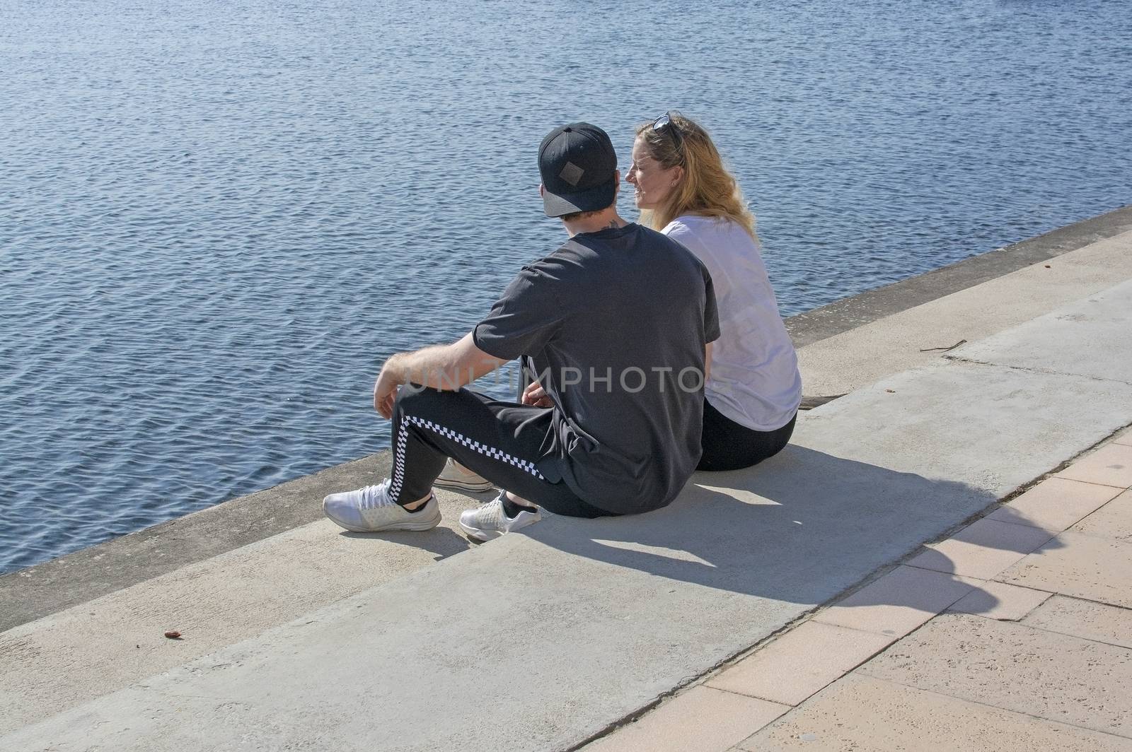 Young couple sit in sunshine watch water glitter on stone pier  by ArtesiaWells