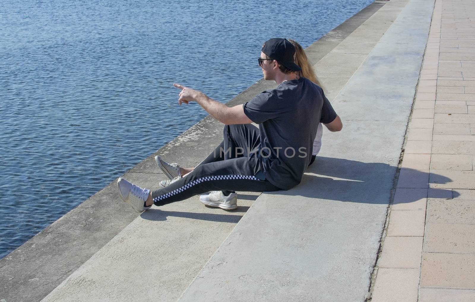 Young couple sit in sunshine watch water glitter on stone pier  by ArtesiaWells