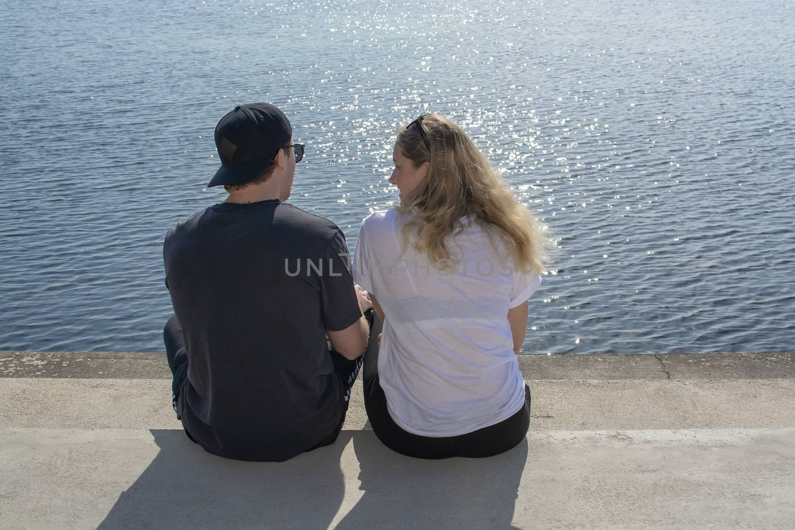 Young couple sit in sunshine watch water glitter on stone pier on a spring day in Mallorca, Spain.