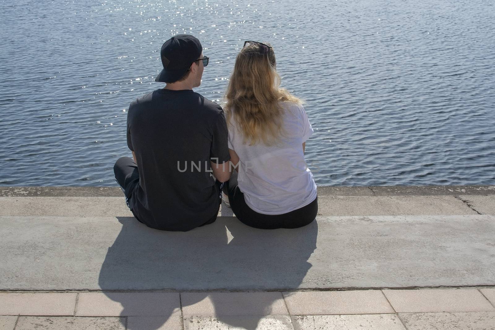 Young couple sit in sunshine watch water glitter on stone pier on a spring day in Mallorca, Spain.