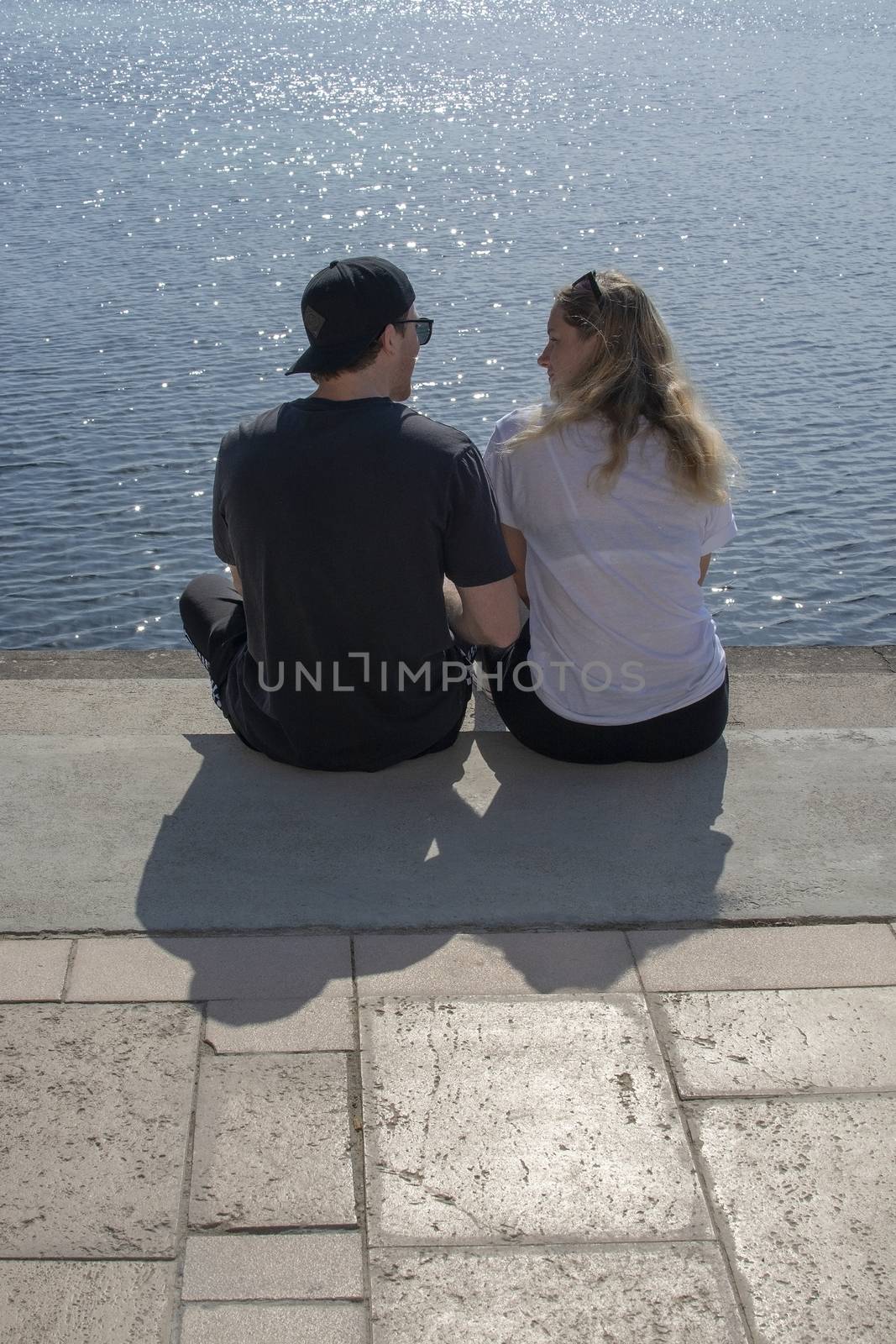 Young couple sit in sunshine watch water glitter on stone pier on a spring day in Mallorca, Spain.