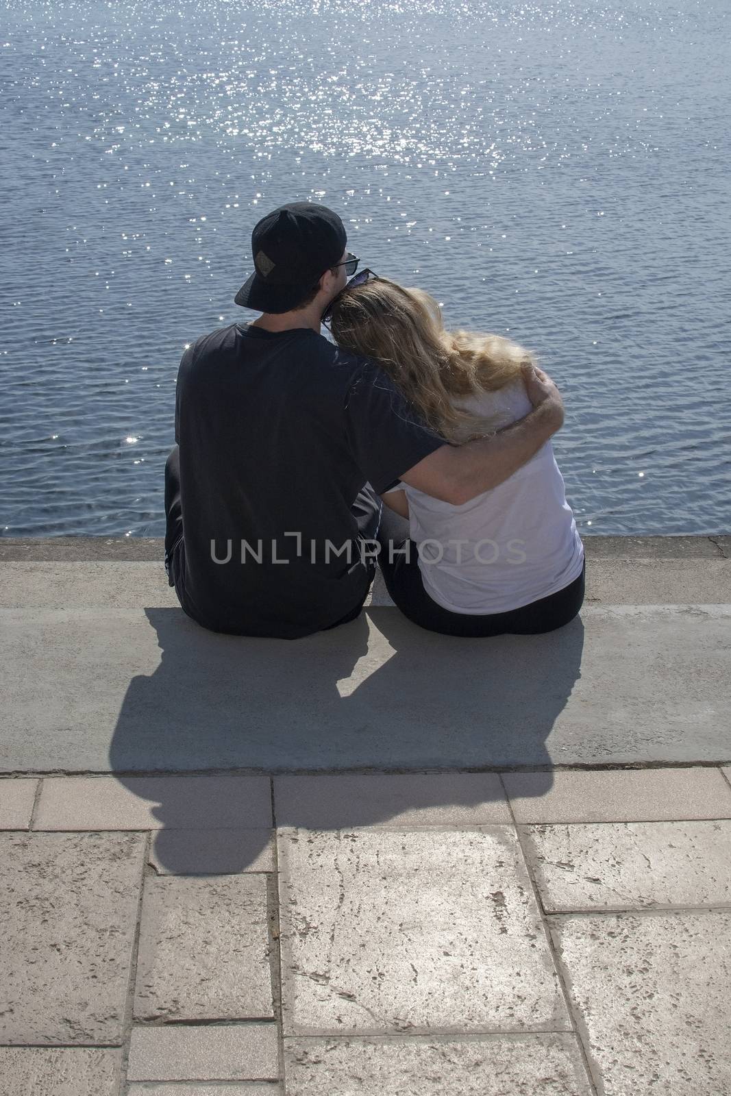 Young couple sit in sunshine watch water glitter on stone pier on a spring day in Mallorca, Spain.