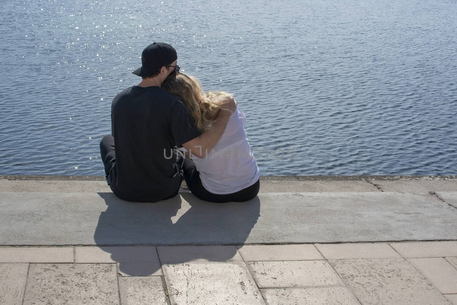 Young couple sit in sunshine watch water glitter on stone pier on a spring day in Mallorca, Spain.