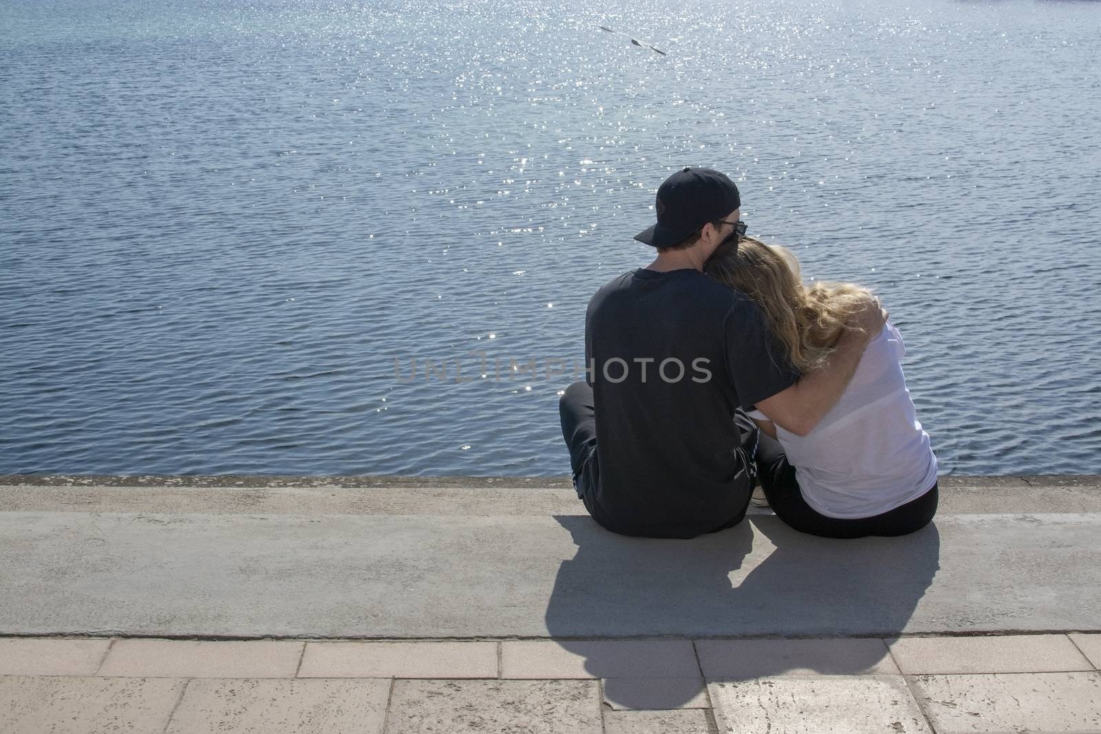 Young couple sit in sunshine watch water glitter on stone pier on a spring day in Mallorca, Spain.