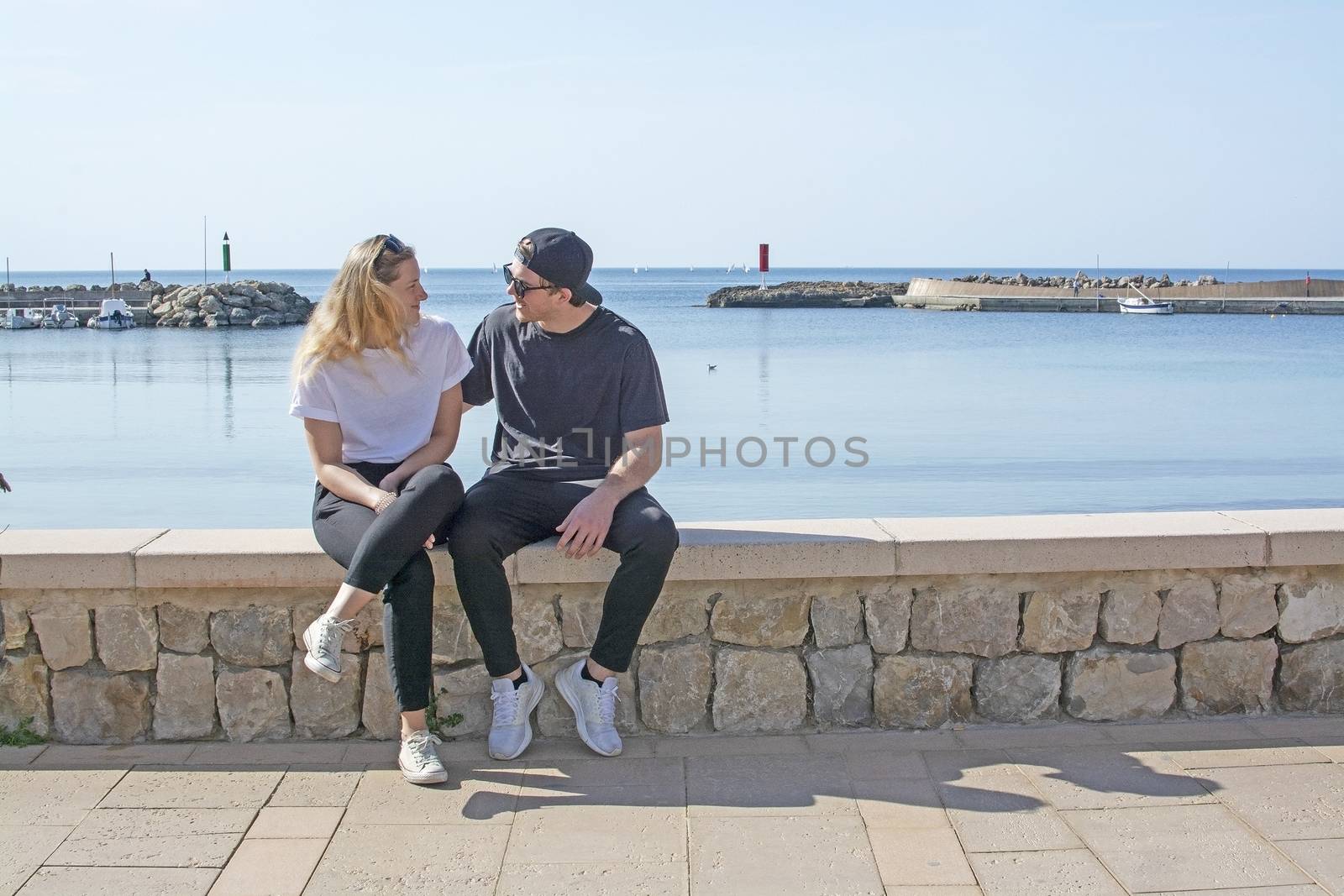 Young natural smiling and casual sporty couple sit on stone wall in sunshine by the ocean in Mallorca, Spain.