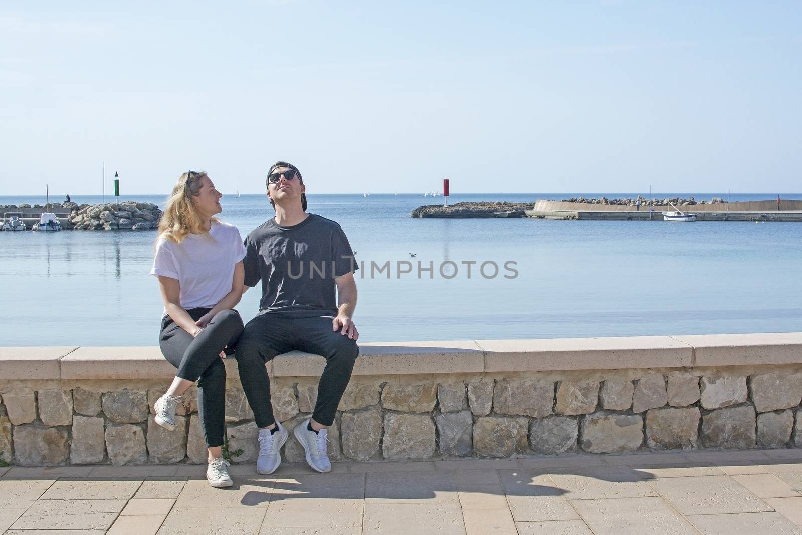 Young natural smiling and casual sporty couple sit on stone wall in sunshine by the ocean in Mallorca, Spain.