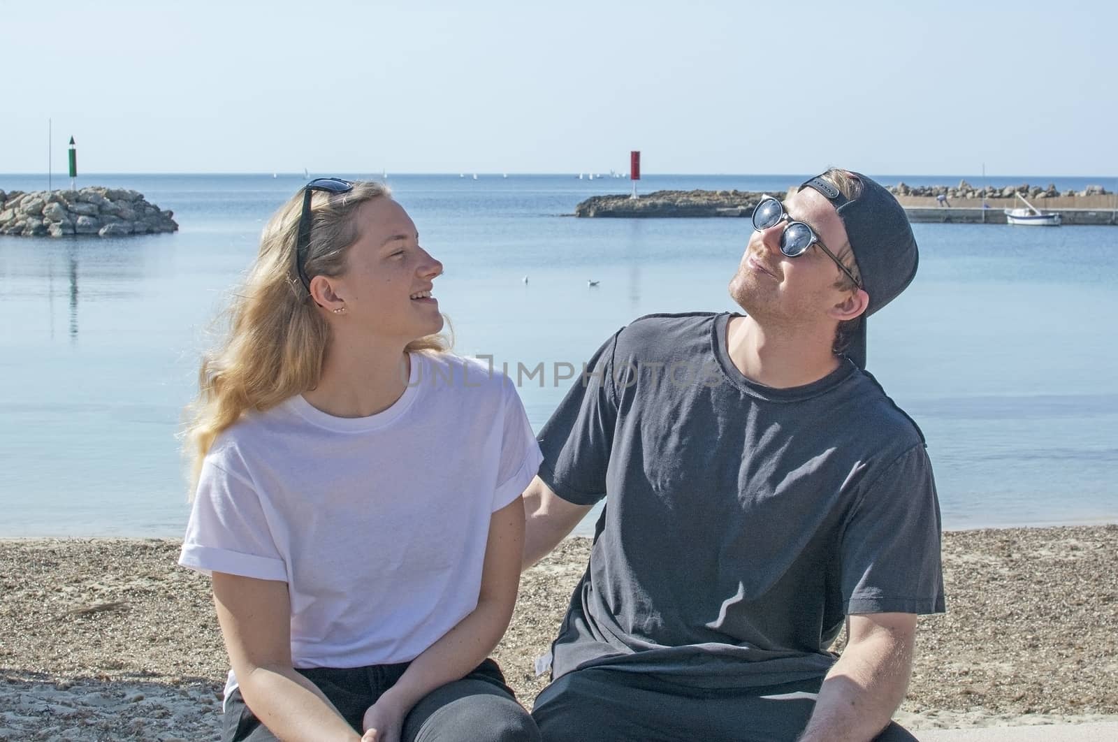 Young natural smiling and casual sporty couple sit on stone wall in sunshine by the ocean in Mallorca, Spain.