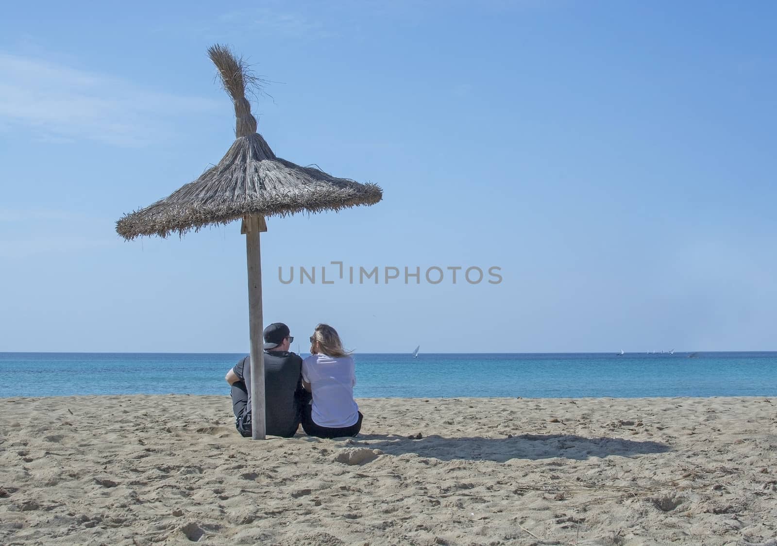 Young natural and casual sporty couple sit on beach under parasol and watch the ocean horizon in Mallorca, Spain.