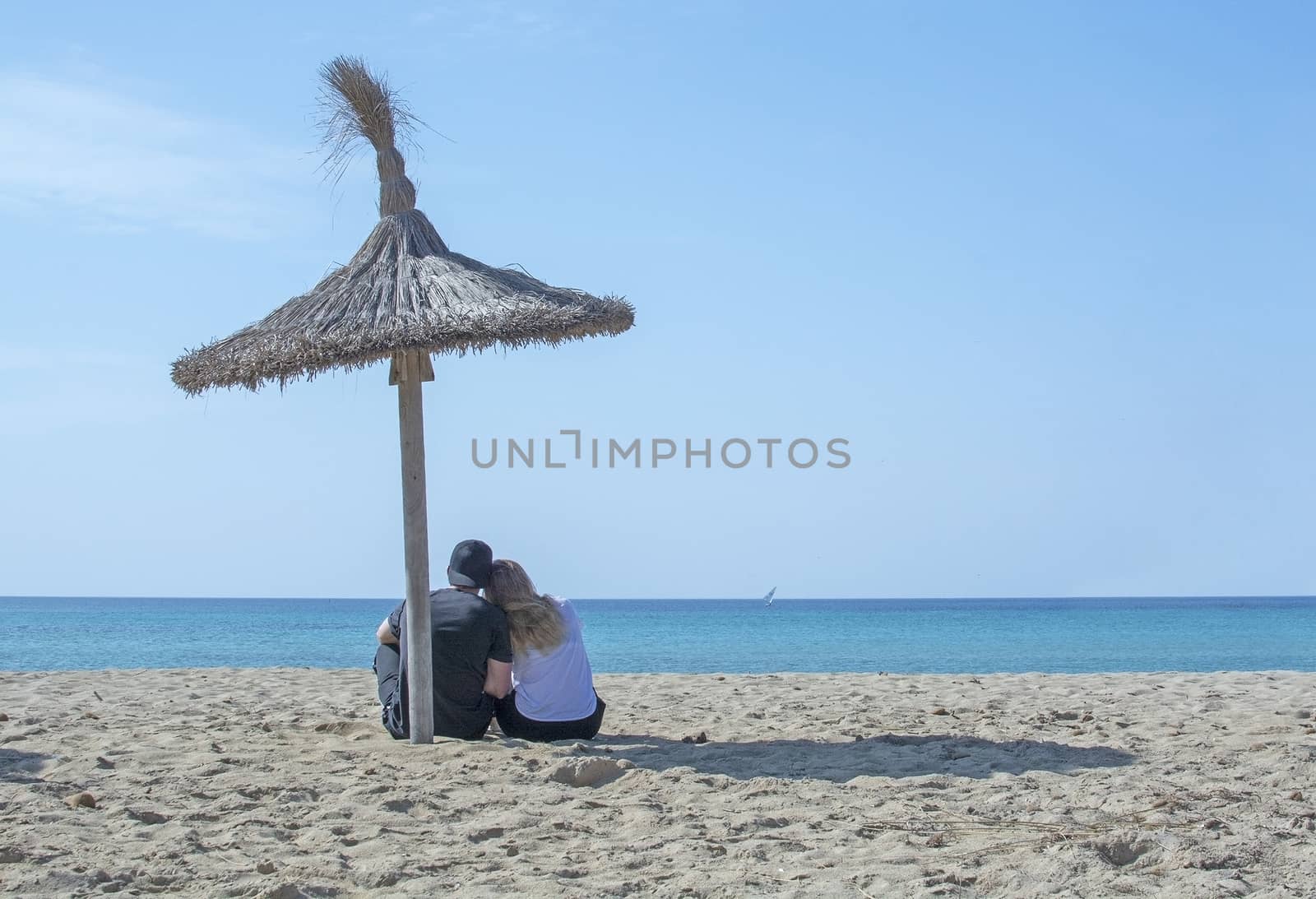 Young couple sit closely together on beach under parasol and watch the ocean horizon in surrealistic romantic pink