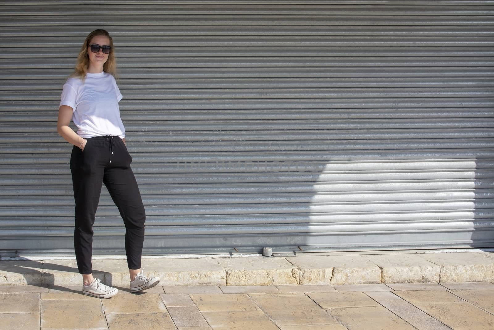 Full body photo of young casual sporty dressed blonde woman with sunglasses in white t-shirt against corrugated iron wall street style