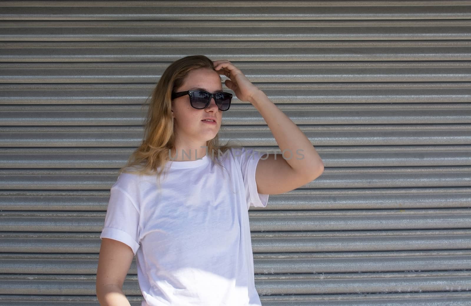 Young casual sporty dressed blonde woman with sunglasses in white t-shirt against corrugated iron wall street style