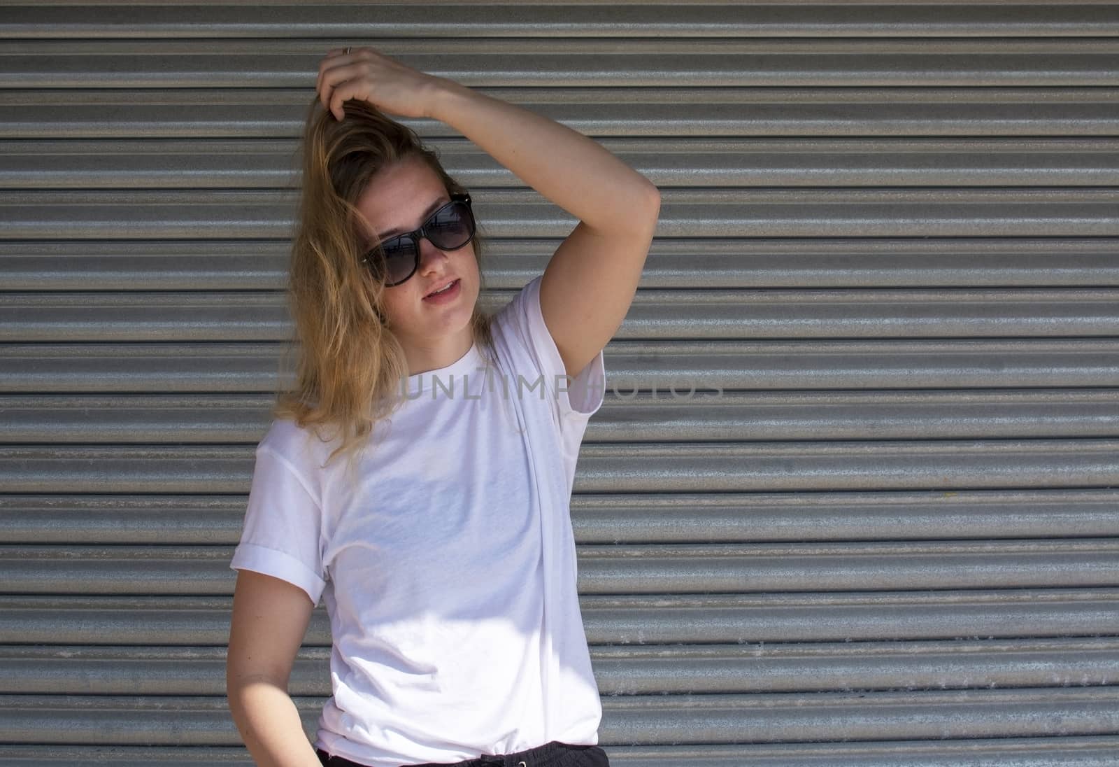 Young casual sporty dressed blonde woman with sunglasses in white t-shirt against corrugated iron wall street style