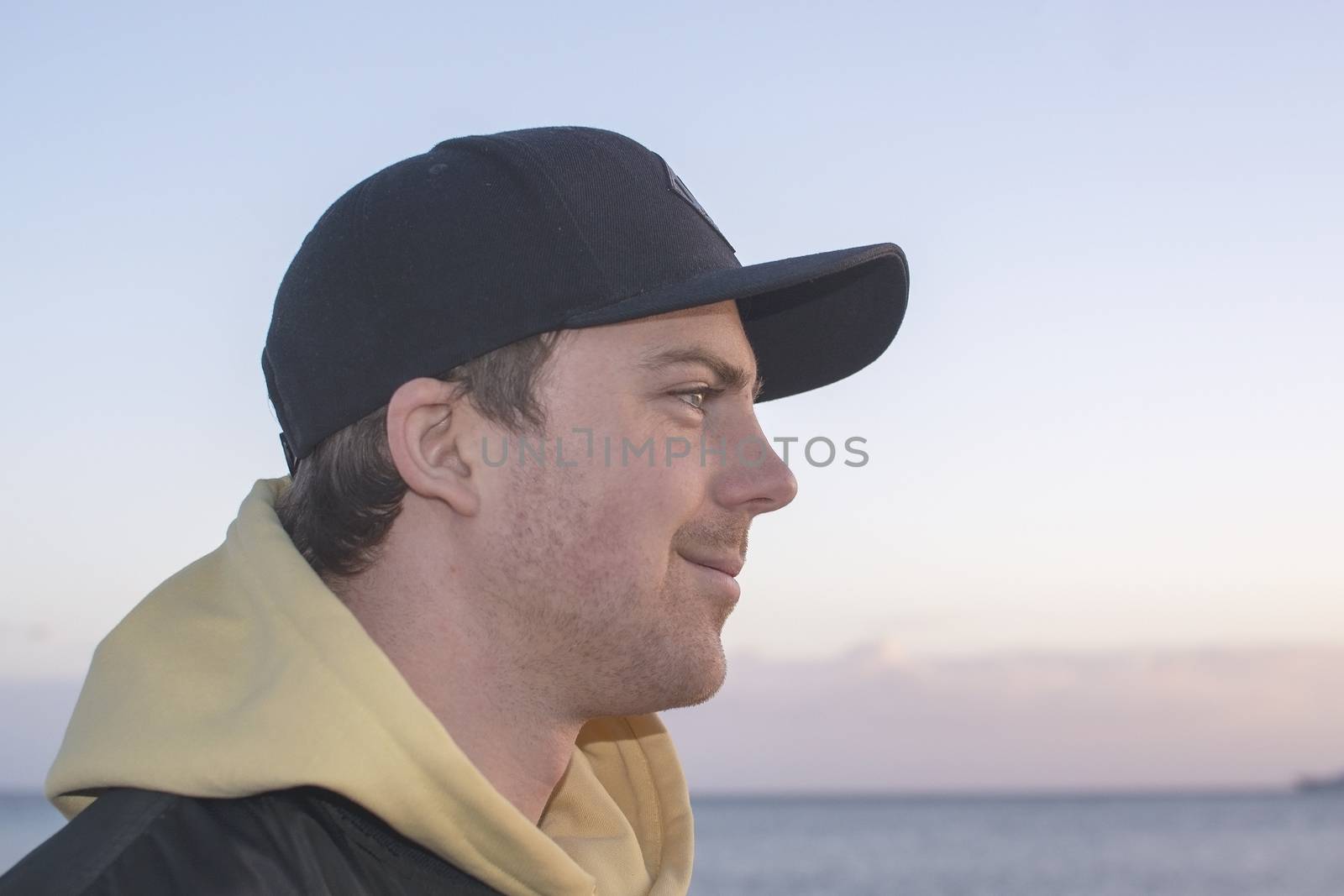 Profile shot of handsome smiling natural and casual looking male in late twenties with hood jacket and cap looks straight forward, ocean horizon behind