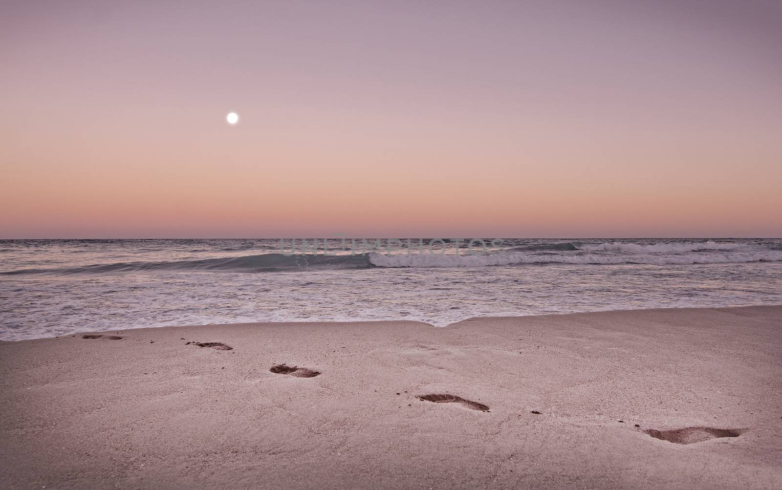 Empty sandy beach with footprints, ocean waves and full moon in expressive dusk orange and purple toned in color Living Coral.