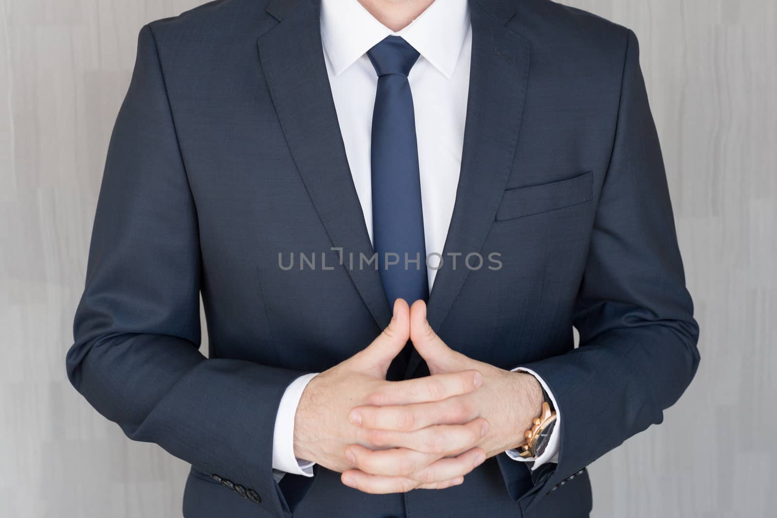 Torso of anonymous businessman standing with hands in lowered steeple, wearing beautiful fashionable classic navy blue suit against grey backgound.