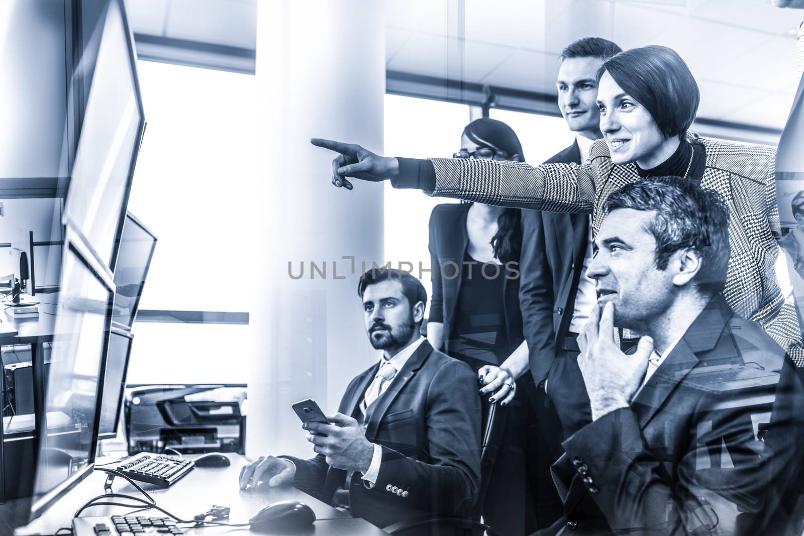 Business people looking at data on multiple computer screens in corporate office. Businesswoman pointing on screen. Business team trading online. Blue toned grayscale image.