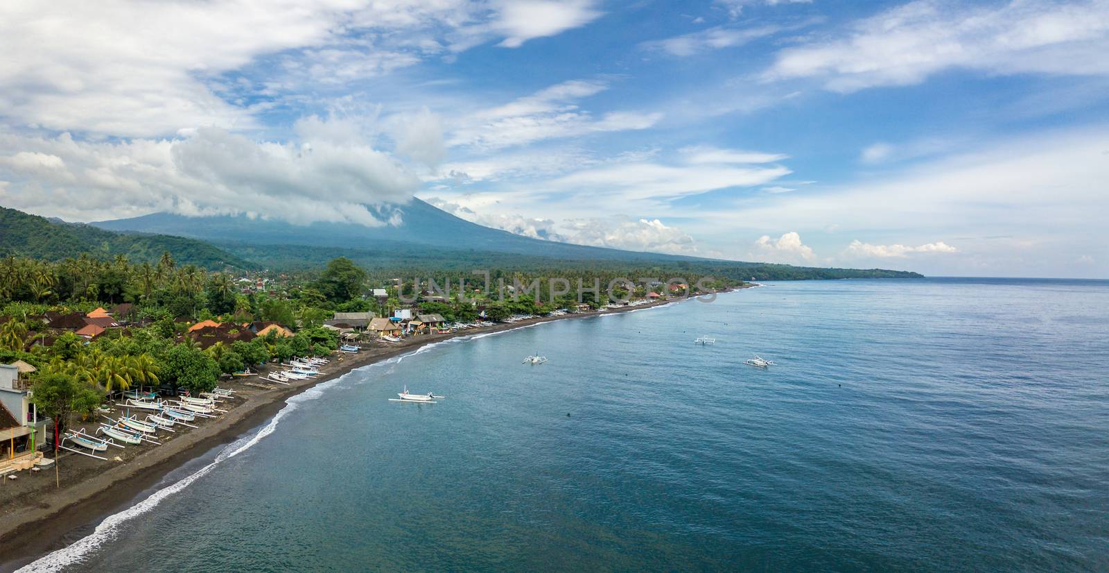 Panoramic aerial view of Amed beach in Bali, Indonesia. Traditional fishing boats called jukung on the black sand beach and Mount Agung volcano in the background, partially covered by clouds.