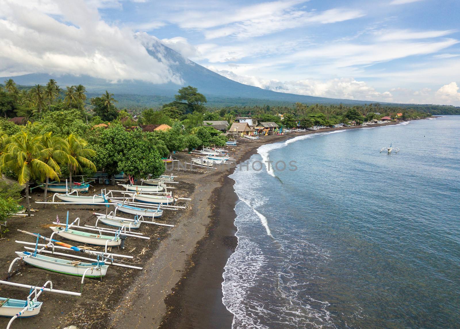 Aerial view of Amed beach in Bali, Indonesia. Traditional fishing boats called jukung on the black sand beach and Mount Agung volcano in the background, partially covered by clouds.