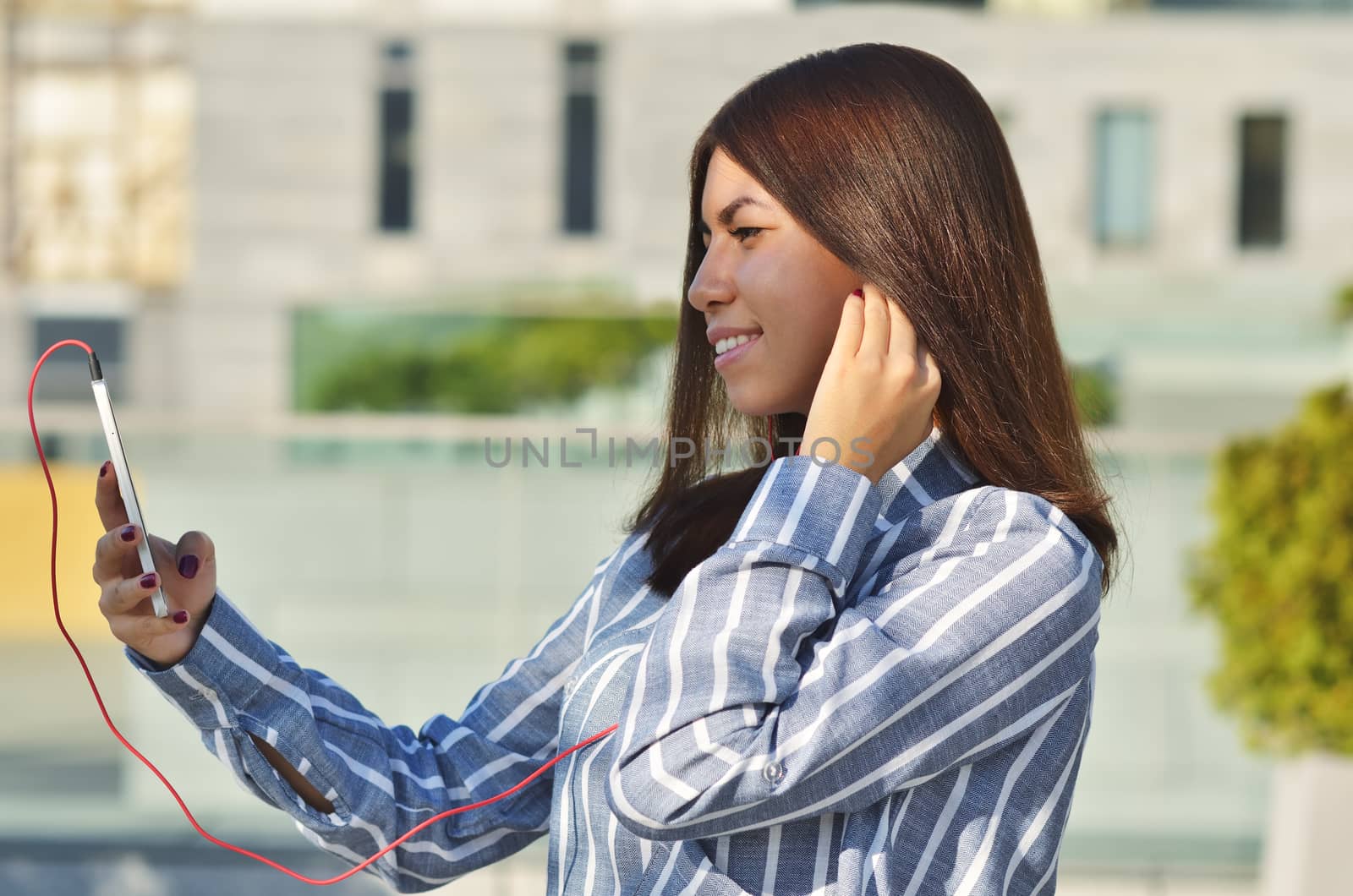 A young girl student of Asian appearance dressed in a striped shirt takes a selfie and listens to music while on the street