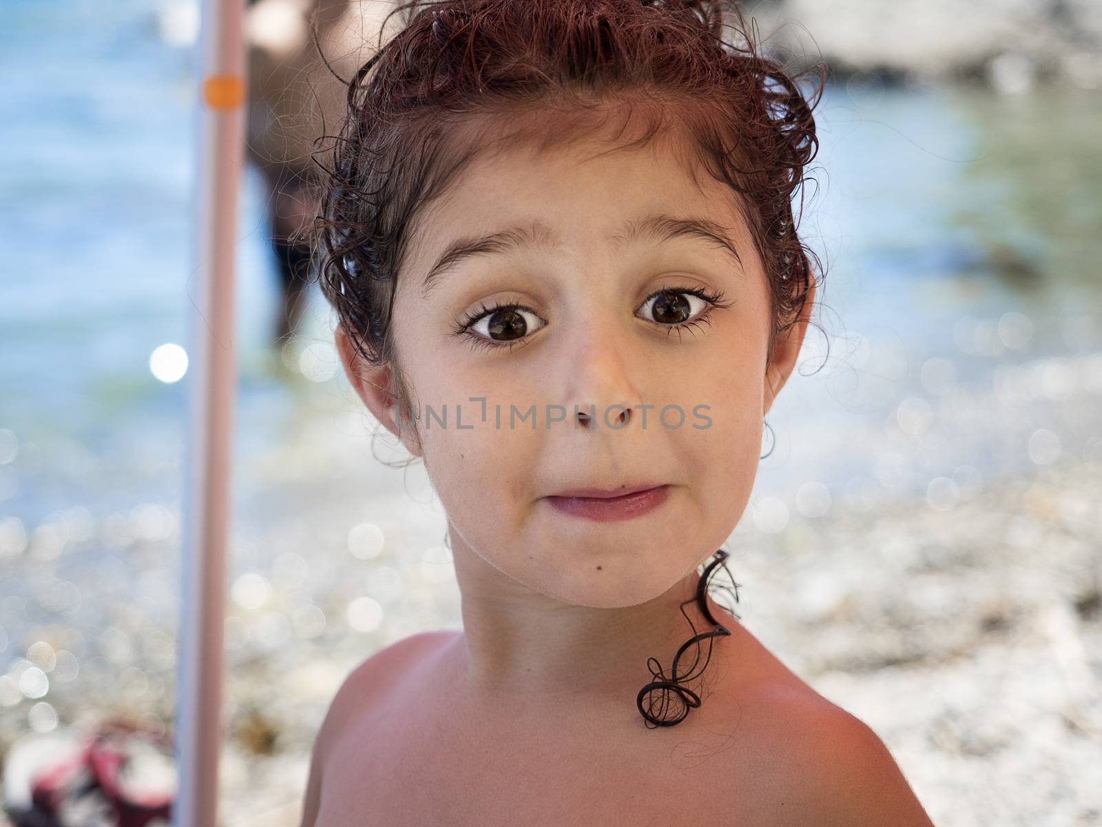 portrait of a kid mocking to the camera, she is shirtless on the beach, her curly hair is wet and she has it picked up, some curls hang down her neck, in the unfocused background you see the seashore