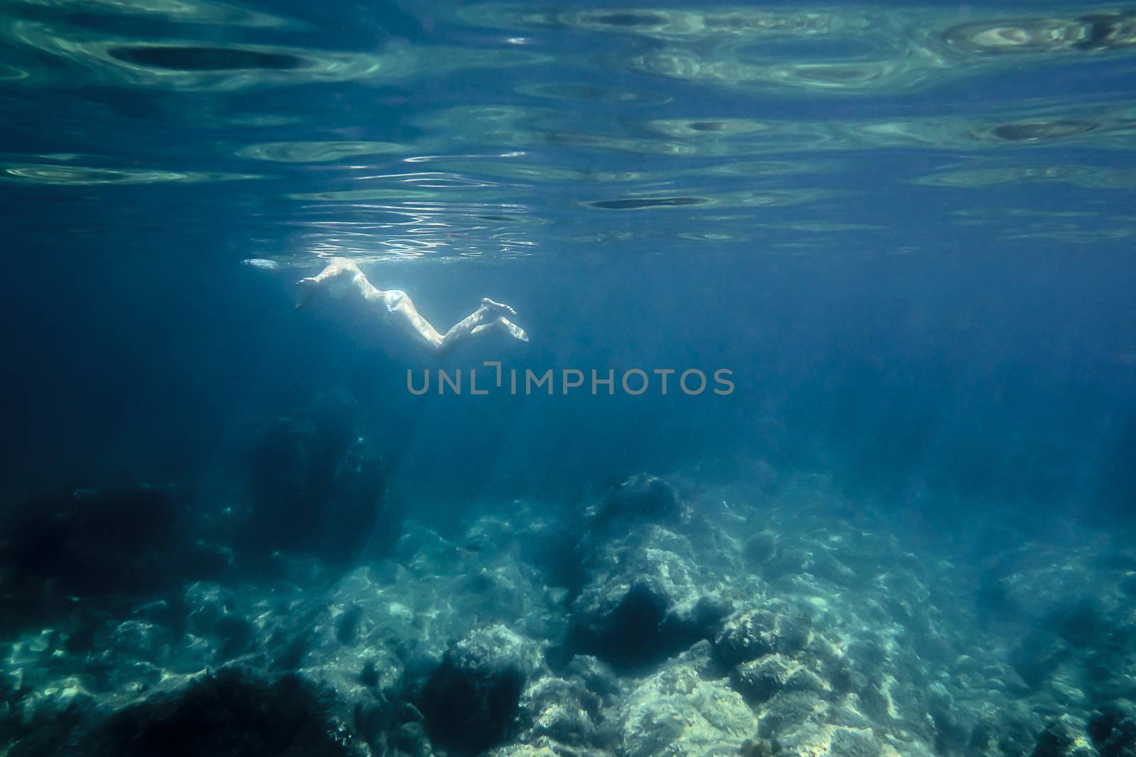 underwater view of a woman relaxing leisurely at sea. The sun reflects on her back and on the curly surface of the sea, also illuminates the rocks of the bottom and light rays are formed in the water