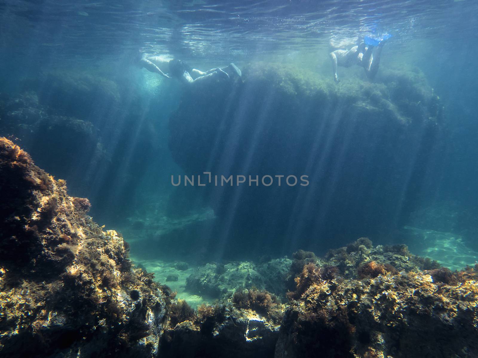 Two people diving near a huge rock seen from under water. Sun rays enter the water illuminating the bottom of sand and rocks