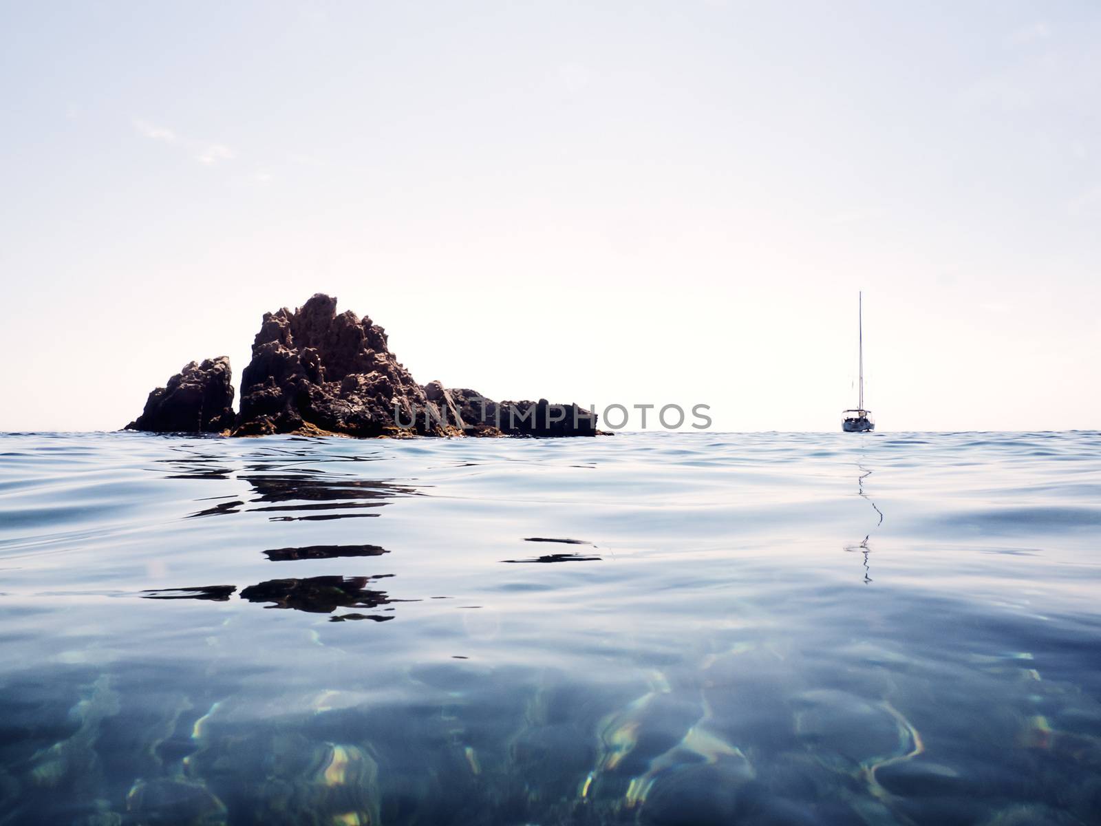 minimalist photograph of a typically Mediterranean marine landscape. The sky is clear, the surface of the sea is calm and there is a sailboat with folded sails near an islet of rocks