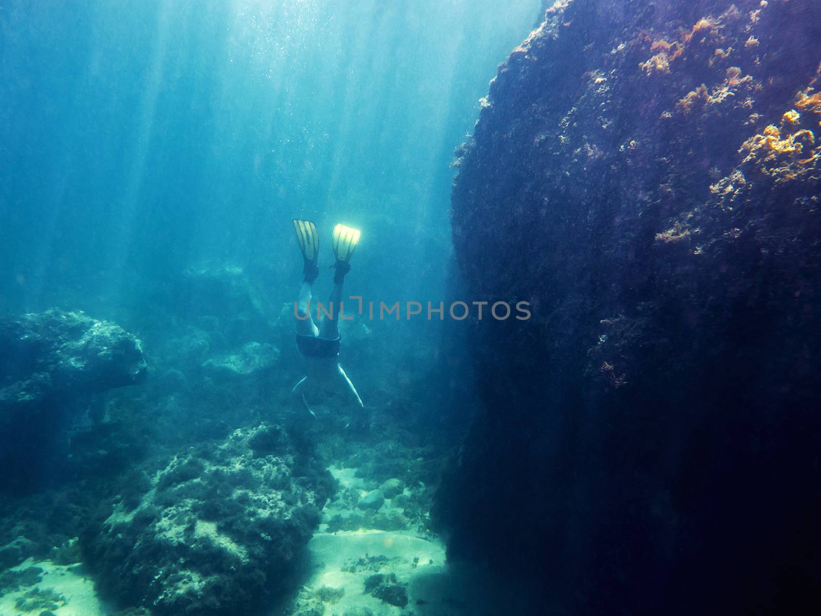 Underwater view of a man diving towards the marine depth with his yellow fins. Sun rays enter the water illuminating the huge rock next to it and the bottom of sand and stones