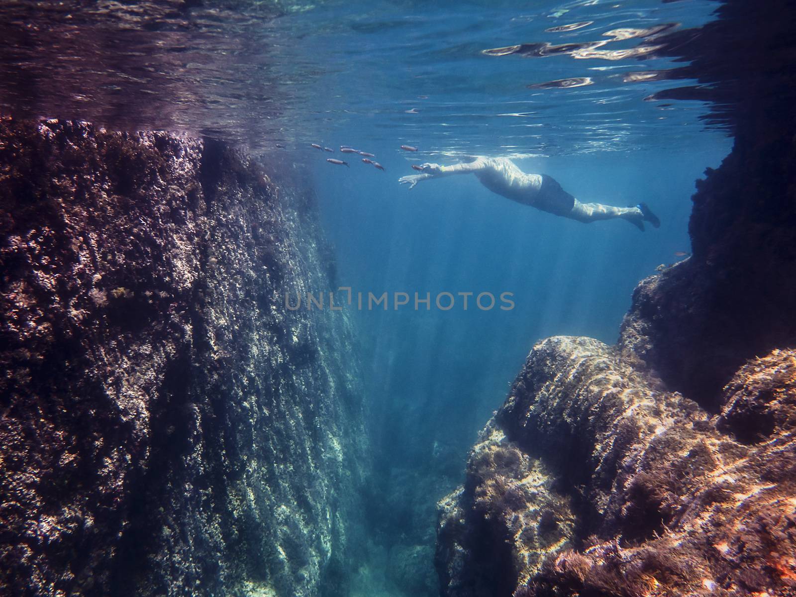 Underwater view of a man swimming placidly in the sea with some fish. Sun rays are drawn in the crystal clear water and illuminate the rocks and curly surface of the blue ocean