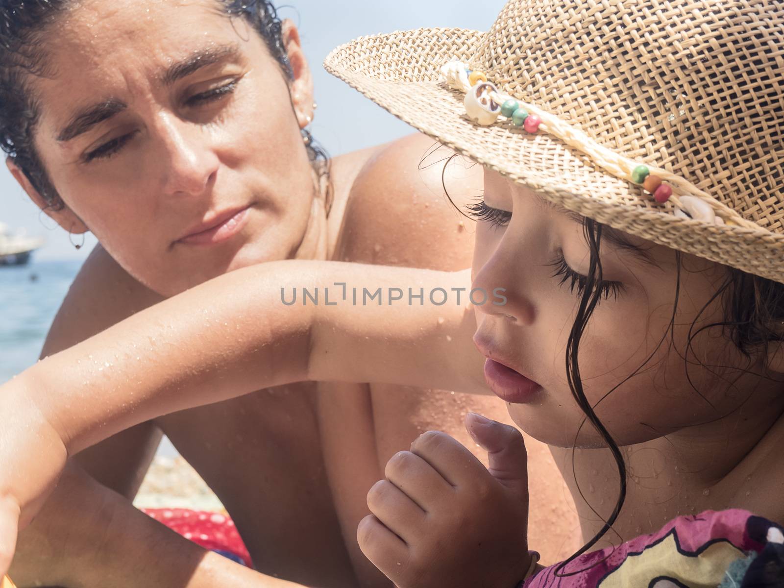 a little girl plays on the beach while her mother looks at her, wears a straw hat with marine ornaments and a lock of curly and wet hair hangs down her face