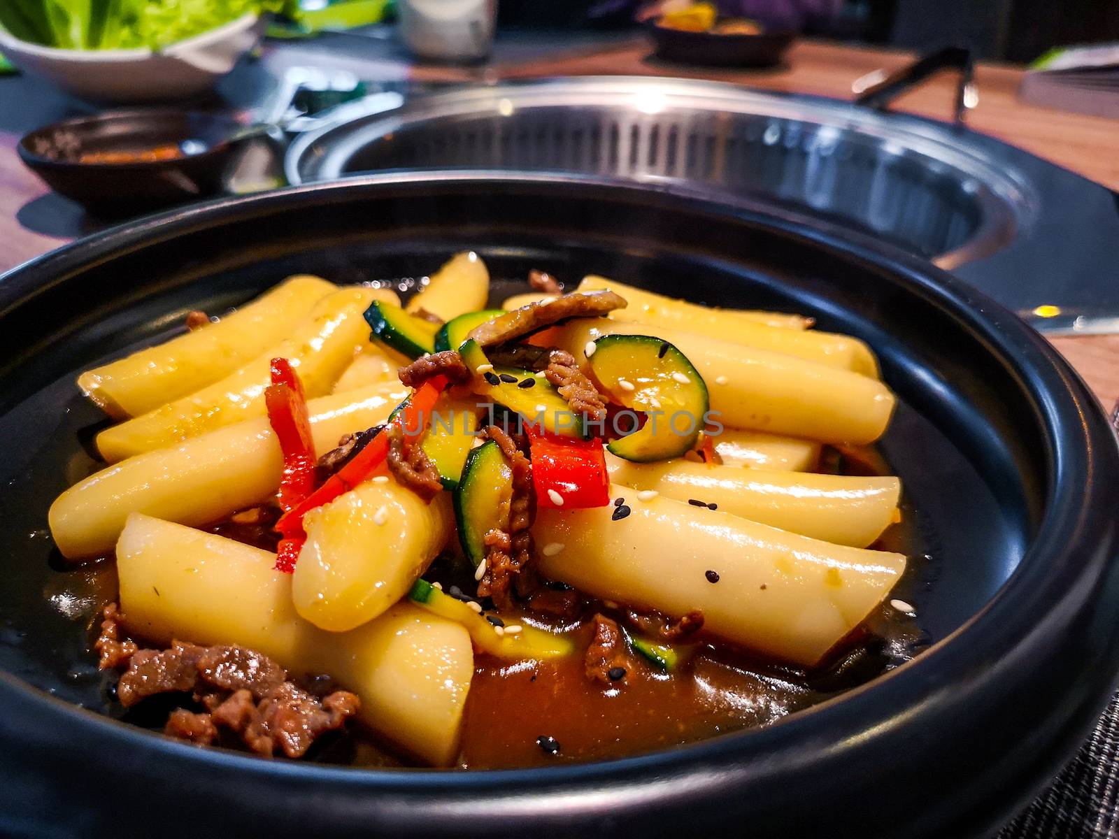 a close up of a Korean Tteokbokki rice dumplings black dish in an korean ethnic restaurant with sweet ganjang soy sauce meat and zucchini bell peppers veggies .