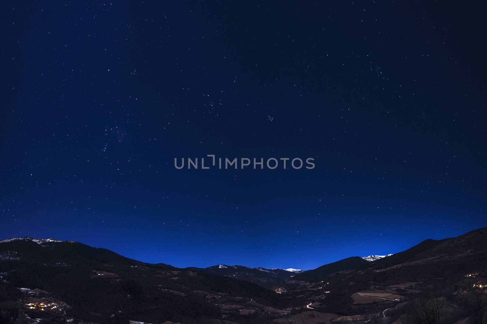 mountain landscape under night sky full of stars, you can see the lights of the houses in the valley and some snowy mountains