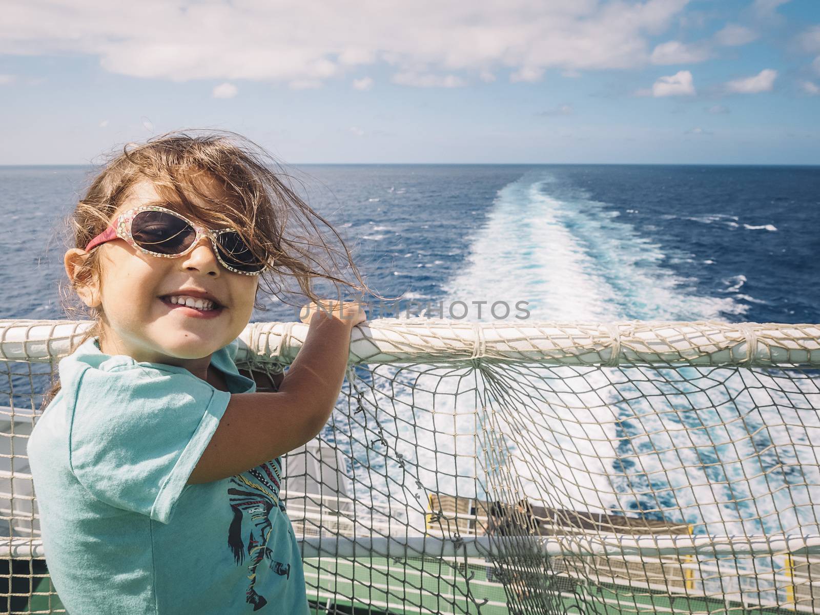 little girl with sunglasses caught at the railing of the cruise ship in which she travels smiles at the camera, the wind moves her hair and the white foam from the ship's wake moves through the ocean