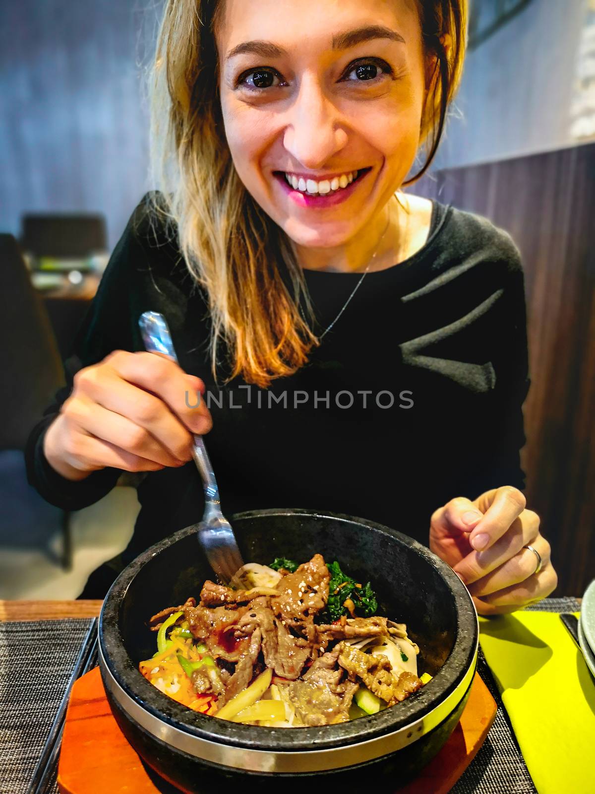korean bibimbap served in hot stone dolsot pot bowl eaten by caucasian smiling woman dining in korean ethnic restaurant by LucaLorenzelli