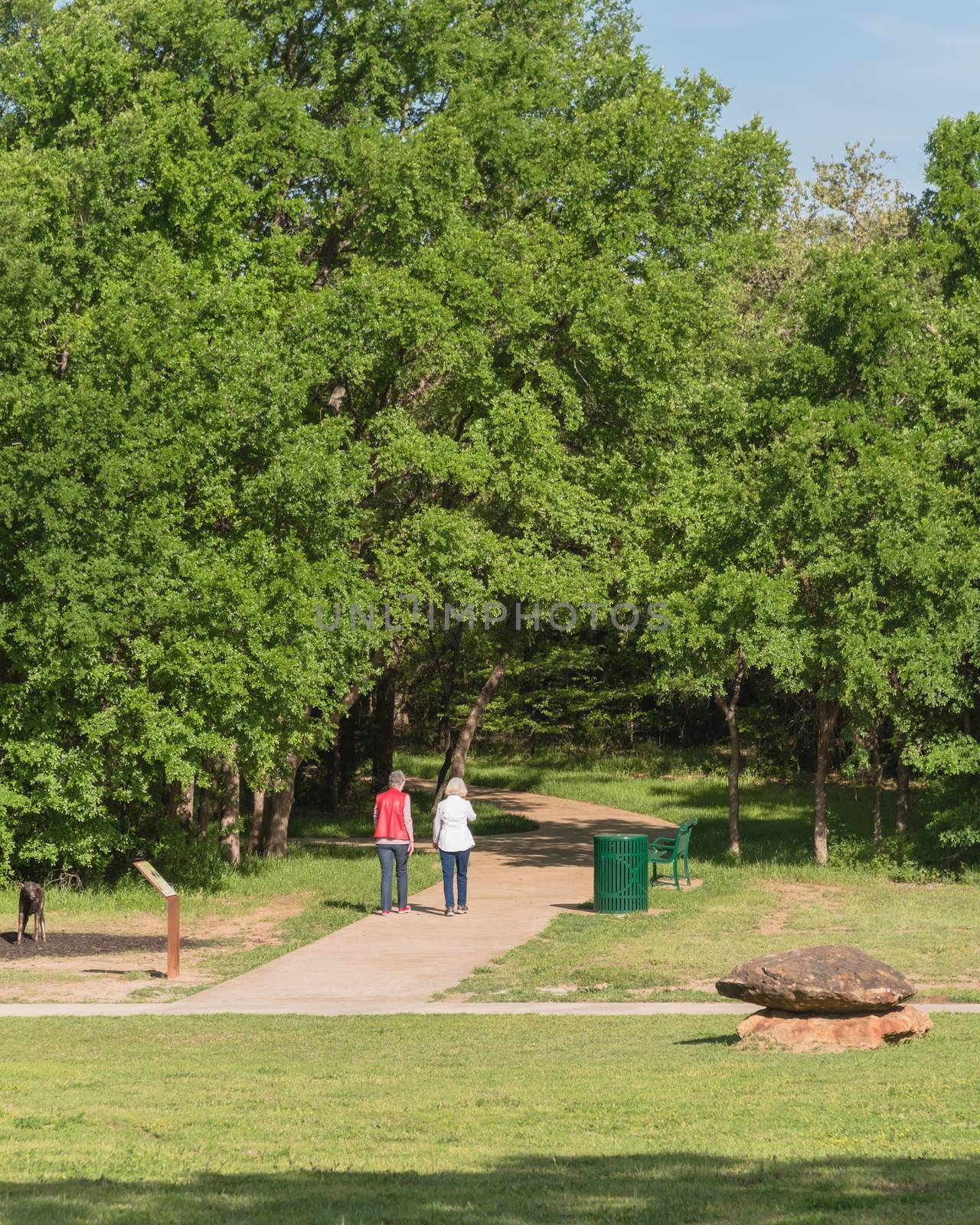 Rear view of senior Caucasian female couple walking in the park. Nature recreation area with trail, trees, bench, trash bin and decorative landscape rock, boulder. Happy retirement concept