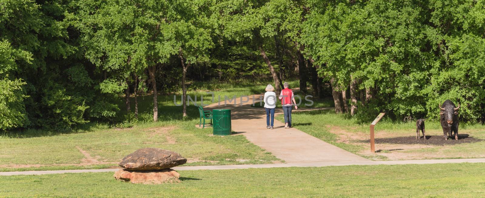 Panorama rear view of senior Caucasian female couple walking in the park. Nature recreation area with trail, trees, bench, trash bin and decorative landscape rock, boulder. Happy retirement concept