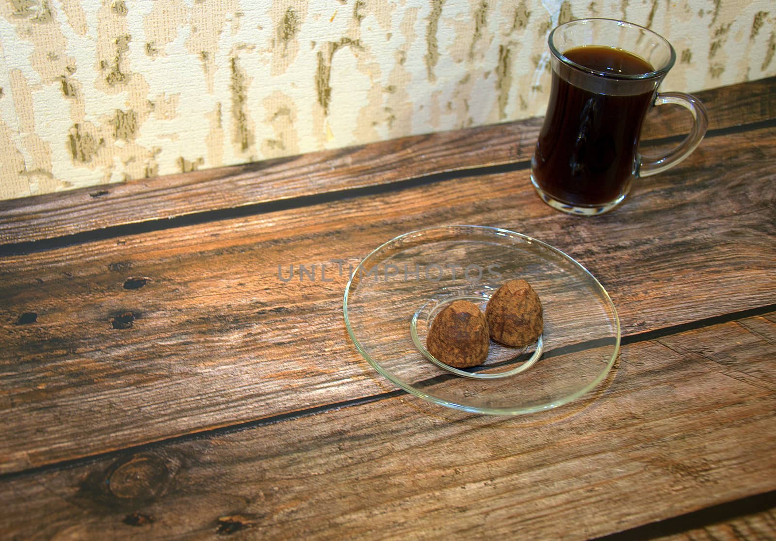 A glass cup of black coffee and a saucer with chocolates on a wooden table.