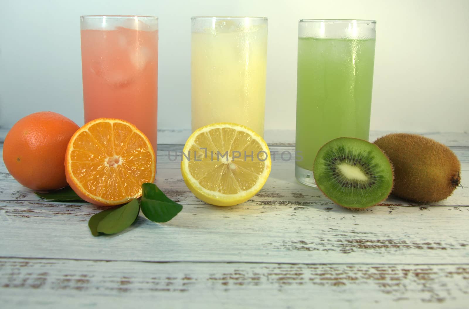 Three glass glasses with a refreshing juice and ice, on a textile stand, whole and sliced half of an orange with leaves, lemon and kiwi, lay on a white wooden table. Close-up.